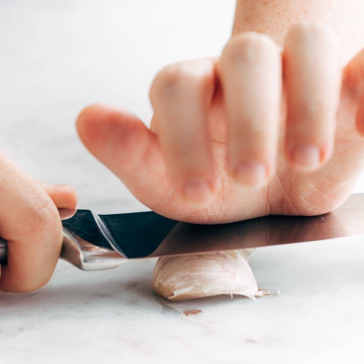 A piece of garlic is being pressed with a knife and pressured by a hand.