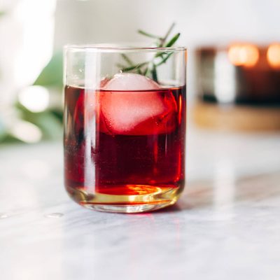A clear cocktail glass with red liquid, a large ice cube, and a sprig of rosemary.